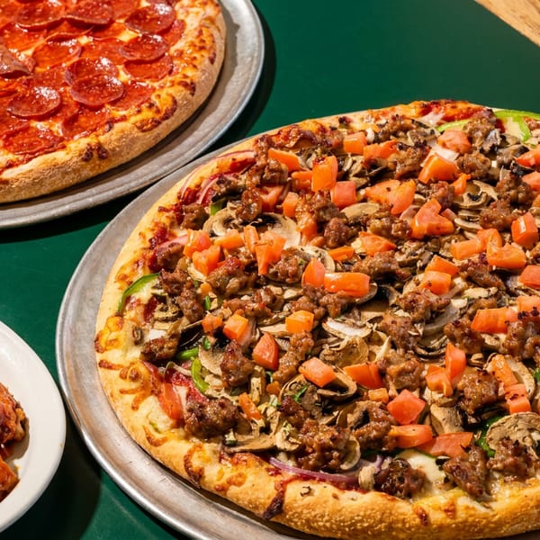 Two pizzas, one pepperoni and one with sausage, mushrooms, and tomatoes, with a side of wings.