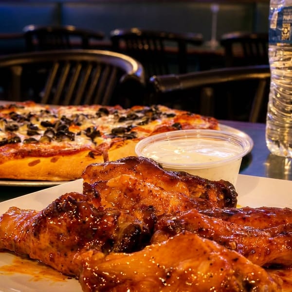 A pizza with black olives and cheese, a plate of BBQ chicken wings, and a water bottle.