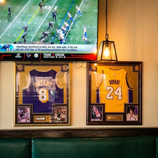 Framed Lakers jerseys of James, Bryant, and West hang below a TV showing a football game.